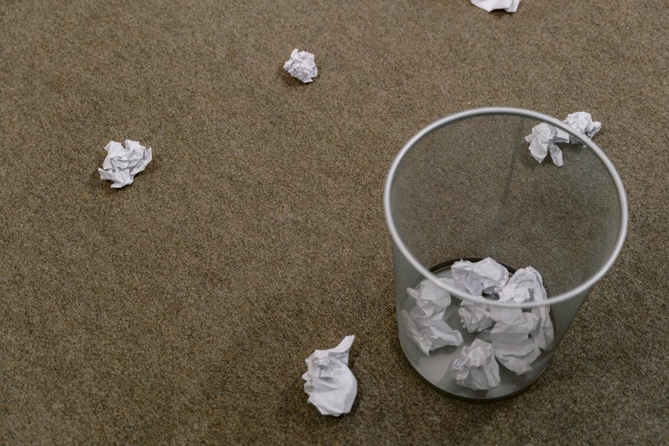 Crumpled paper scattered around a trash bin on a carpeted floor, indoors scene.